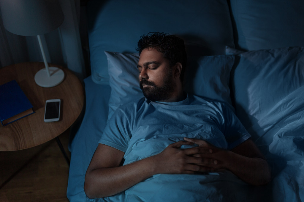 man peacefully sleeping in a blue silk bed with nightstand on the side